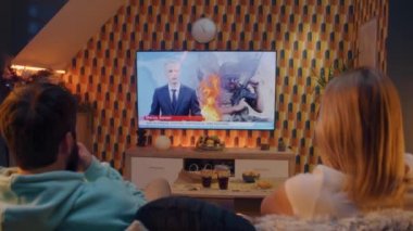 Oturma odasındaki kanepede oturan çift ellerinde abur cuburlar, konuşuyorlar, Ukrayna 'daki savaş ve Rus istilası hakkında TV haberleri izliyorlar. Kadın ve erkek geceyi evde geçiriyorlar. TV canlı yayın.