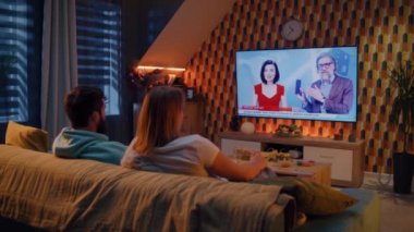 Oturma odasındaki koltukta oturan, abur cubur ve gazozla konuşan, TV haberleri izleyen, yeni telefon veya dijital ürün sunumu yapan, hafta sonunu rahat bir evde birlikte geçiren çift. TV canlı yayın.