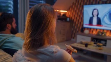 Oturma odasındaki kanepede oturan çift, son dakika haberleri izliyor, pizza ve cips yiyor. Karı koca hafta sonları evde dinleniyor ve dinleniyorlar. TV canlı yayın.