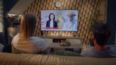 Oturma odasında birlikte oturan çift, modern iç mekanla birlikte televizyon kanalında Covid 19 salgınının yeni dalgasını izliyorlar. Erkek ve kadın hafta sonunu televizyon tıp programında geçiriyorlar.