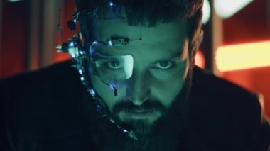 Esmer, sakallı, fütürist kulaklık takan, tek gözlü, mikrofon ve kulaklık takan adam kameraya yoğun bir yüz ifadesiyle bakıyor. Arka planda neon ışıkları var. Siber Punk.