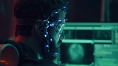 Fütürist kulaklıklı ve mikrofon programlı, birden fazla bilgisayar ekranı kullanan sakallı bir gencin yan görüntüsü. Cyperpunk tarzı. Bilim kurgu geçmişi. Neon ışıkları.
