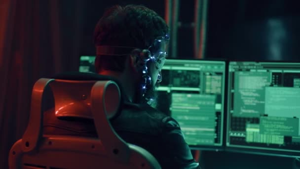 Ledライト付きの未来的なヘッドセットを身に着けているブルネットの男のバックビューは プログラムを添付し 複数のコンピュータ画面を使用してコードを開発します サイパーパンク風 背景にSf ネオンライト — ストック動画