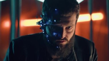 Esmer, sakallı, fütürist kulaklık takan, tek gözlü, mikrofon ve kulaklık takan adam kameraya yoğun bir yüz ifadesiyle bakıyor. Arka planda neon ışıkları var. Siber Punk.