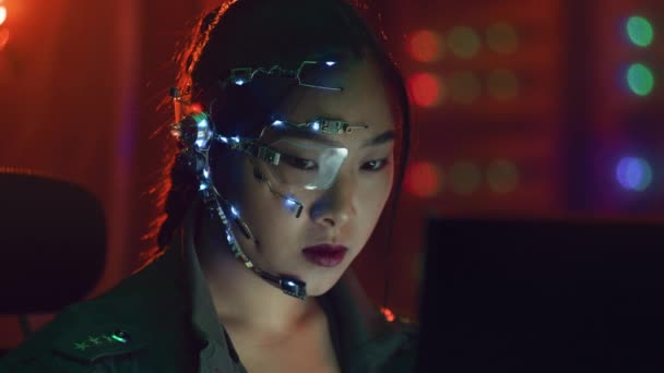 サイバーパンクの女の子は 赤いネオンでコンピュータ上で動作します 未来的な片目のメガネとマイクを持つアジアの少女 サイバーとサイエンスの背景 — ストック動画