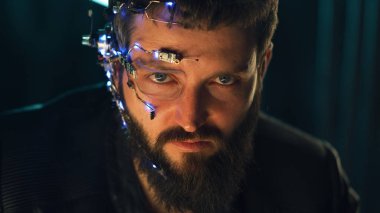 Esmer, sakallı, fütürist kulaklık takan, tek gözlü, mikrofon ve kulaklık takan adam kameraya yoğun bir yüz ifadesiyle bakıyor. Arka planda neon ışıkları var. Siber Punk.
