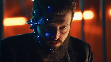 Esmer, sakallı, fütürist kulaklık takan, tek gözlü, mikrofon ve kulaklık takan adam kameraya yoğun bir yüz ifadesiyle bakıyor. Arka planda neon ışıkları var. Siber Punk.