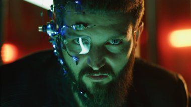 Esmer, sakallı, fütürist kulaklık takan, tek gözlü, mikrofon ve kulaklık takan adam kameraya yoğun bir yüz ifadesiyle bakıyor. Arka planda neon ışıkları var. Siber Punk.