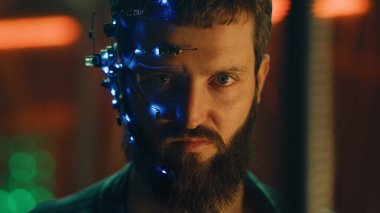 Esmer, sakallı, fütürist kulaklık takan, tek gözlü, mikrofon ve kulaklık takan adam kameraya yoğun bir yüz ifadesiyle bakıyor. Arka planda neon ışıkları var. Siber Punk.