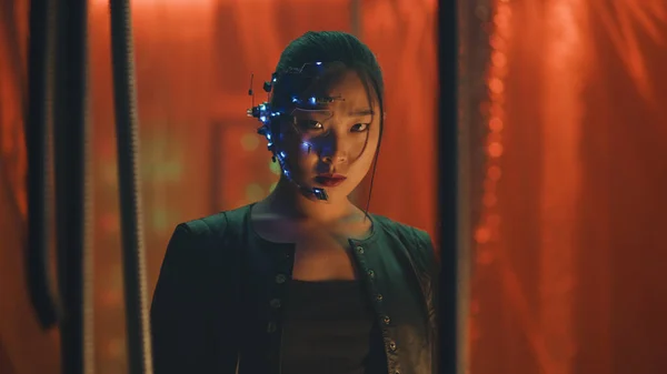 Cyberpunk girl in a black leather jacket looks fiercely at the camera. Asian girl with one-eyed glasses and headset trapped. Black thick rods hanging to prevent her from escaping.