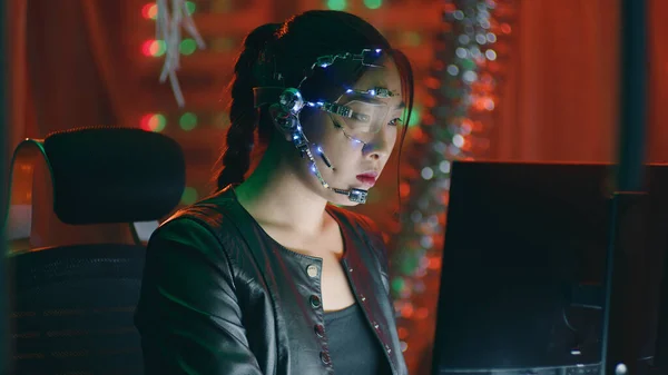 Focused cyberpunk girl in black leather jacket works on the computer. Wearing a headset with white LED lights and microphone. Cyberpunk background with neon lights.