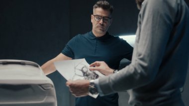 Deneyimli araba tasarımcıları ve geliştiriciler prototip araba heykelinin önünde dururken arabanın tasarımını tartışırlar. Yüksek teknoloji ürünü bir otomobil imalat şirketinin kalem çizimlerini kullanarak..