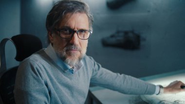 Siyah çerçeveli gözlüklü ve sakallı yaşlı bir otomotiv tasarımcısının yakın çekimi işi durdurur ve kameraya bakar. Yüksek teknolojili yenilikçi bir laboratuvarda prototip model arabanın çizimleri..