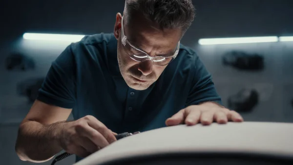 Experienced car designer works closely on the prototype car model in a high tech car manufacturing company. Wearing safety goggles using a handheld rotary tool for cutting edge.