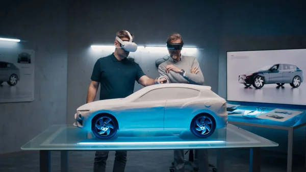 Automotive professional engineers with VR headset and joysticks talk about vehicle production while standing infront of a prototypecar model in a high tech office. Car design analysis and improvement.