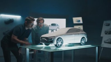 Kıdemli otomotiv mühendisleri, yüksek teknolojili bir ofiste konsept araba modeli bir prototiple çalışıyorlar. Duvara büyük bir LED ekran ve araba modeli çizimleri monte ediyorlar. Model mükemmelleştiriliyor ve tasarım düzeltmeleri yapılıyor.