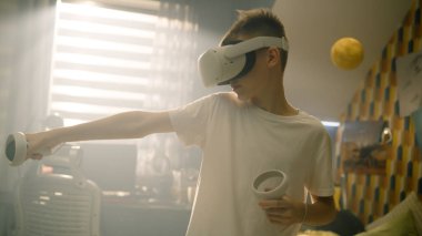 VR kulaklıklı ve elinde joysticklerle heyecanlı bir genç. Genç çocuk sanal gerçeklik kulaklığı kullanıyor ve odasında oyunlar oynuyor..