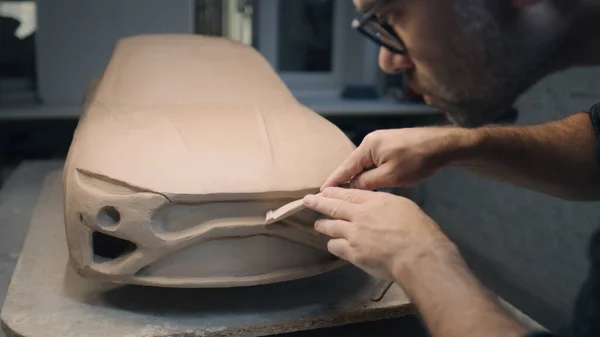 Hands of a male designer works on the sculpture of a car using sculpting tools. Smooths out the surface of the unfinished model in a workshop. Automotive industry.