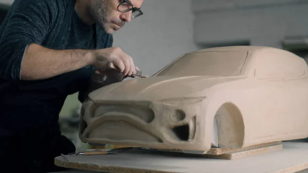 Experienced car designer works on the prototype model of a car using wooden shaping tools to smooth out the bonnet surface. Unfinished car model placed on a wooden table.