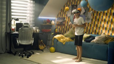 Genç çocuk sanal gerçeklik kulaklığı takarak ve elinde joysticklerle bir boks oyunu oynuyor. Saldırı ve savunma hareketleri yapıyor. Genç çocuk odasında VR kulaklık kullanıyor ve oyunlar oynuyor..