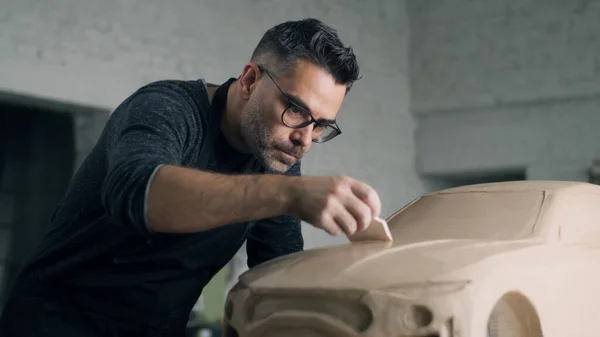 Experienced automotive designer with an apron works on the clay sculpture of the eco-friendly car dragging using wooden sculpting tool to carve the design details. Hand made car sculpture made of clay