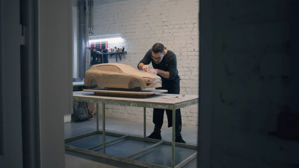 Male designer with an apron sculpting prototype model of a car made of clay placed on a wooden table in a workshop. Uses sculpting tools to create details design.