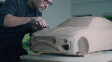Deneyimli araba tasarımcısı, kaput yüzeyini düzeltmek için ahşap şekillendirme araçları kullanan bir arabanın prototip modeli üzerinde çalışıyor. Tamamlanmamış bir araba modeli ahşap bir masaya yerleştirilmiş..