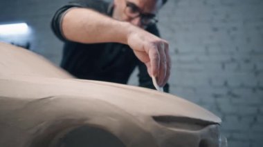 Üst düzey araba tasarımcısı, heykel aletleri kullanan bir prototip araba modelinin kil heykeli üzerinde çalışıyor. Siyah çerçeveli gözlüklü araba tasarımcısı tasarımın ayrıntıları üzerinde çalışıyor. Arabanın prototip modeli..