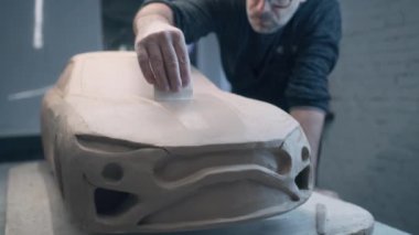 Deneyimli araba tasarımcısı, yontma aletleri kullanılarak ham kilden yapılmış bir prototip araba modeli üzerinde çalışıyor. Çerçeveli gözlüklü araba tasarımcısı araba kaputunun yüzeyini düzleştirmek için yakından çalışıyor.