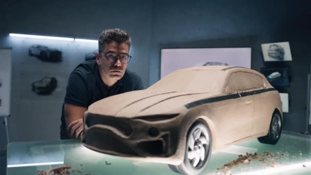 プラスチック粘土から未来的な車のモデルの近くに立っている中年の男性の自動車デザイナーは カメラを見て 新世代電気自動車の将来設計 現代のカーデザインスタジオでの仕事 — ストック動画