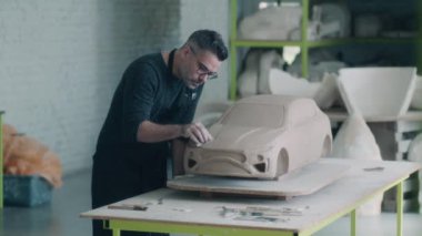 Önlüklü deneyimli bir araba tasarımcısı, yüzeyi düzleştirmek ve tasarımda ayrıntılar yaratmak için ahşap şekillendirme aracı kullanarak arabanın heykeli üzerinde çalışıyor. El yapımı araba heykeli kilden yapılmış..