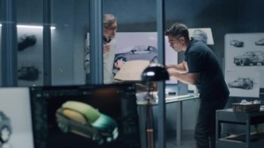 İki erkek otomotiv tasarımcısı elektrikli arabanın fütüristik plastik kil modelini tırmıkla yontuyorlar. Modern araba tasarım stüdyosunda çalışıyorum. 3B modelleme projeleri için yazılım içeren bilgisayar ekranı.
