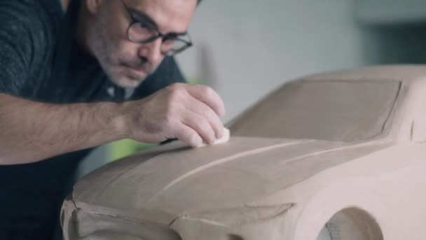 経験豊富な自動車設計者は 車の彫刻プロトタイプの表面を滑らかにするために 木製の成形ツール全体にドラッグします 車の粘土彫刻にエプロンをつけたデザイナー — ストック動画