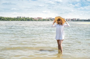 Beyaz bluzlu ve hasır şapkalı bir kadın sıcak bir yaz gününde gölde yürüyor.