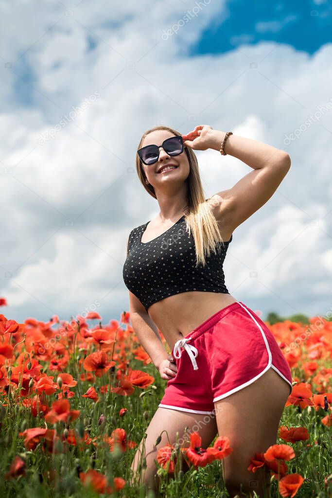 young woman in sunglasses and black top standing at the field of poppy flowers. hot summer day