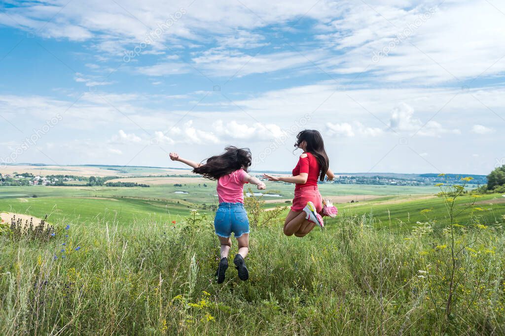 two happy slim smiling young women jumping together on rural field on the blue sky
