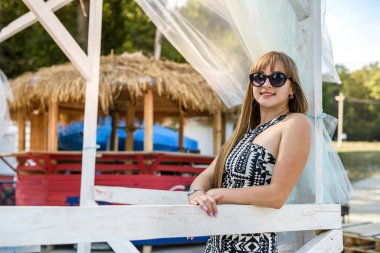 Genç bayan yazın beyaz çardakta gölün yanında poz veriyor.