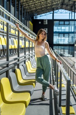 Spor giyimli genç, güzel, zayıf bir kadın stadyumdaki koltukların yanında rahatlıyor.