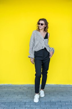 Açık havada, sarı duvarın yanında güneş gözlüklü gri örülü süveter giyen pozitif kadın modeli.
