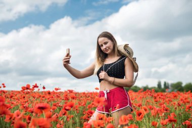 Siyah bluzlu ve şapkalı güzel Ukraynalı kırmızı haşhaş tarlasında güneşli günün tadını çıkar. 