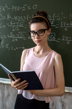 Genç ciddi bayan öğretmen üniversitede matematik formüllerini karatahtanın yanında açıklıyor.  
