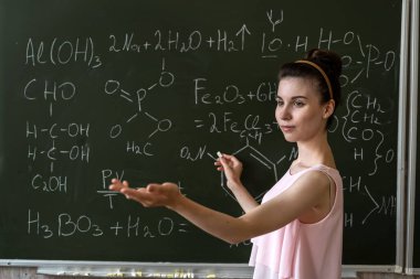 Bayan öğretmen kimyasal formülleri karatahta ve eğitim konseptinin yanında açıklıyor.