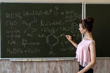 Bayan öğretmen kimyasal formülleri karatahta ve eğitim konseptinin yanında açıklıyor.