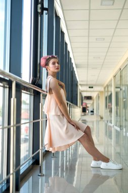 Pembe elbiseli ve beyaz ayakkabılı bir kadın koridorda poz veriyor.