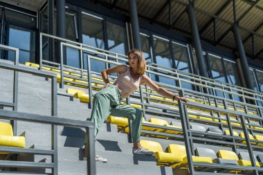 Spor giyimli genç, güzel, zayıf bir kadın stadyumdaki koltukların yanında rahatlıyor.