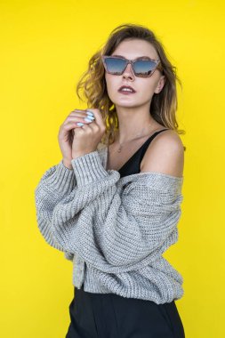 Açık havada, sarı duvarın yanında güneş gözlüklü gri örülü süveter giyen pozitif kadın modeli.