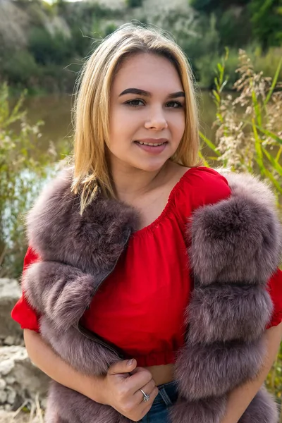 毛皮のコートを着た美しい女の子と暖かい秋の日に湖がポーズをとる薄い赤いTシャツとショートパンツ — ストック写真