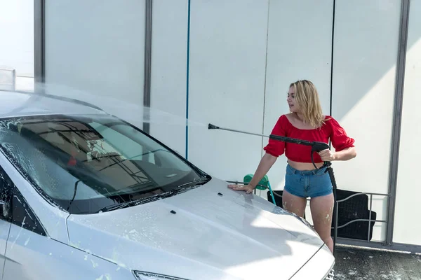 Kadın oto yıkama istasyonunda arabasının köpüğünü yüksek basınçlı hortumla yıkıyor.