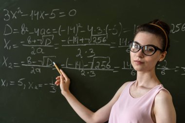 Genç ciddi bayan öğretmen üniversitede matematik formüllerini karatahtanın yanında açıklıyor.  