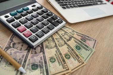 Tahta bir masada dizüstü bilgisayar, hesap makinesi, dolar banknotları, kalem var. İş konsepti